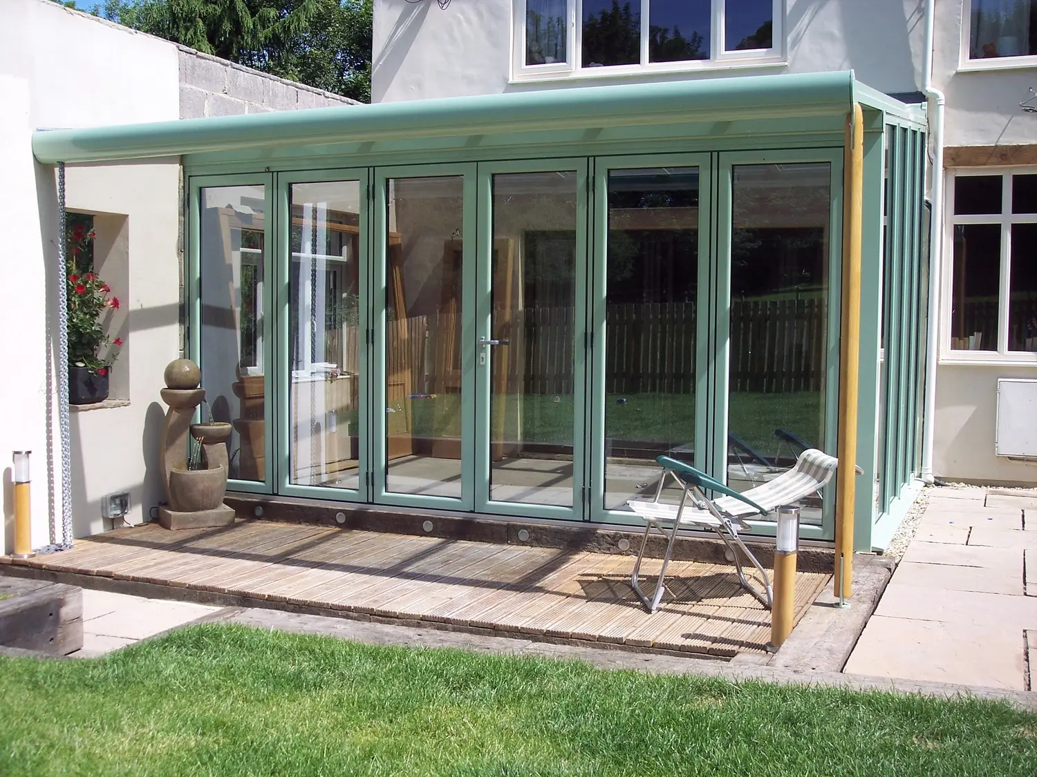A six-panel bifold door run in sage green with a matching green roof, folding open onto a decked patio