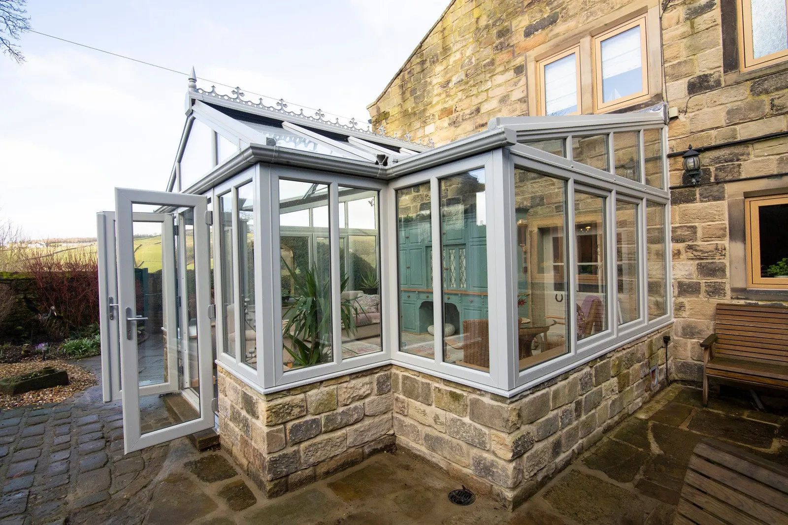 A white-framed conservatory with a glazed roof against a stone house, seen from the patio