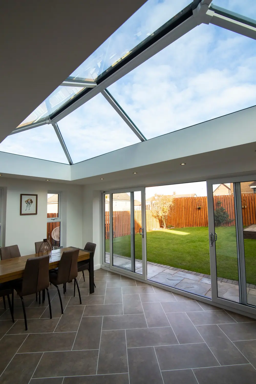 White sliding patio doors along a dining room, beneath a large roof lantern, opening onto a lawn