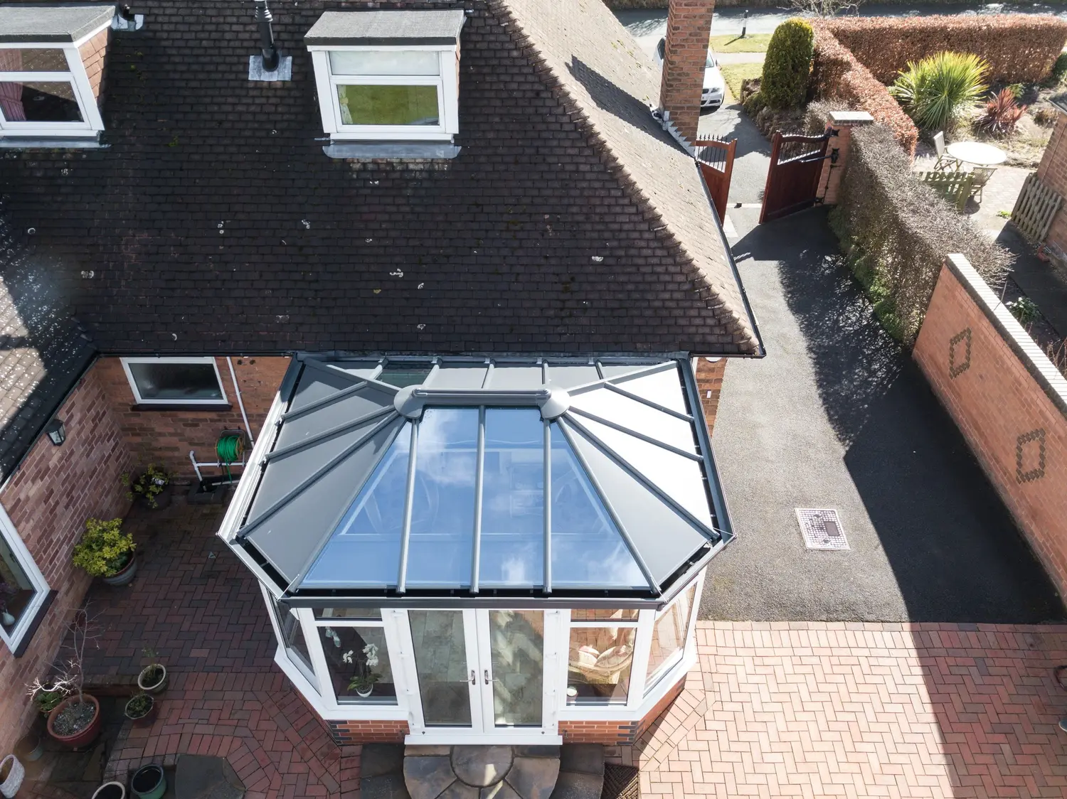 A conservatory with a dark glazed roof on a brick house, seen from above with the driveway and road beyond